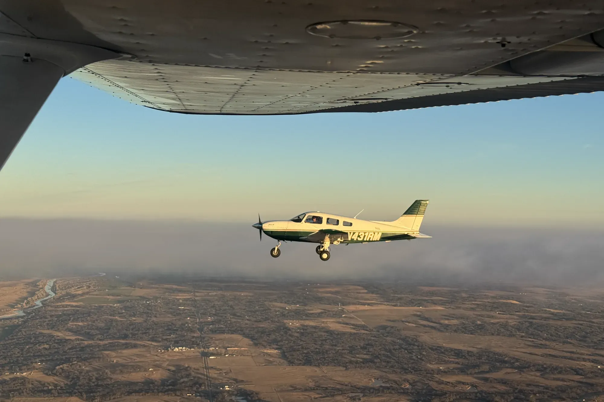 N431RM in flight over farmland and a winding river in low evening sun, photographed from beneath the wing of another aircraft.
