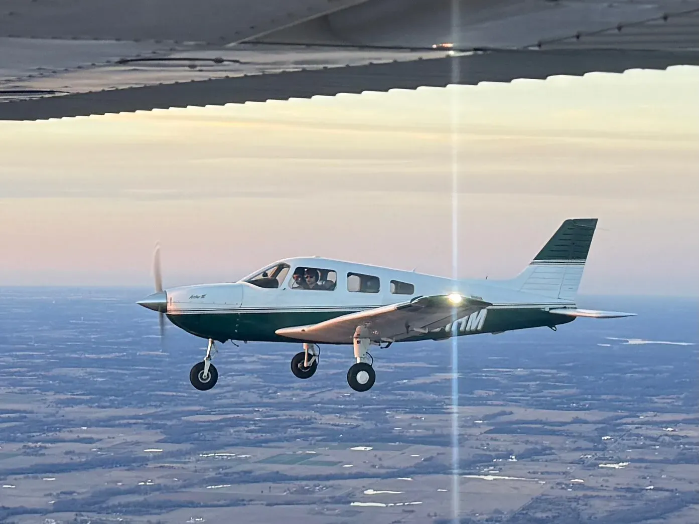 N431RM in flight in the evening, seen from the left side beneath the wing of another aircraft, with two people in the cockpit and farmland far below.